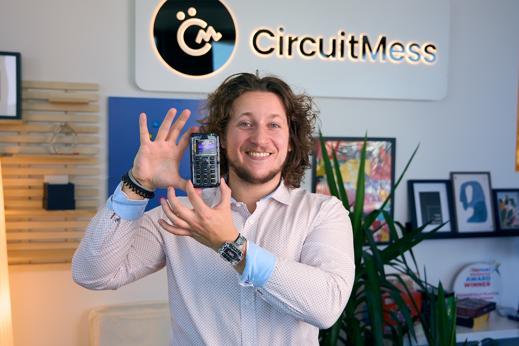 Albert Gajšak holding up a MAKERphone with both hands, smiling, under the CircuitMess logo on the office wall.
