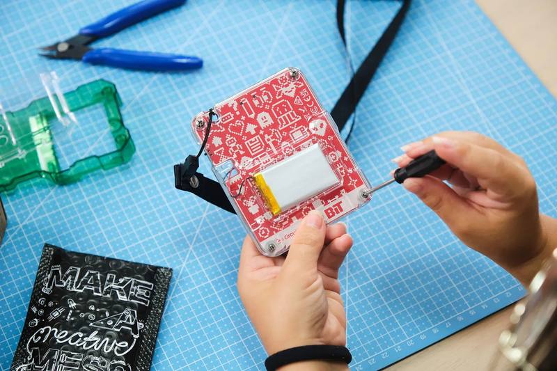 Overhead view of two hands screwing the back plate onto a red Bit 2.0 console on a blue cutting mat, with the battery and rear board visible.