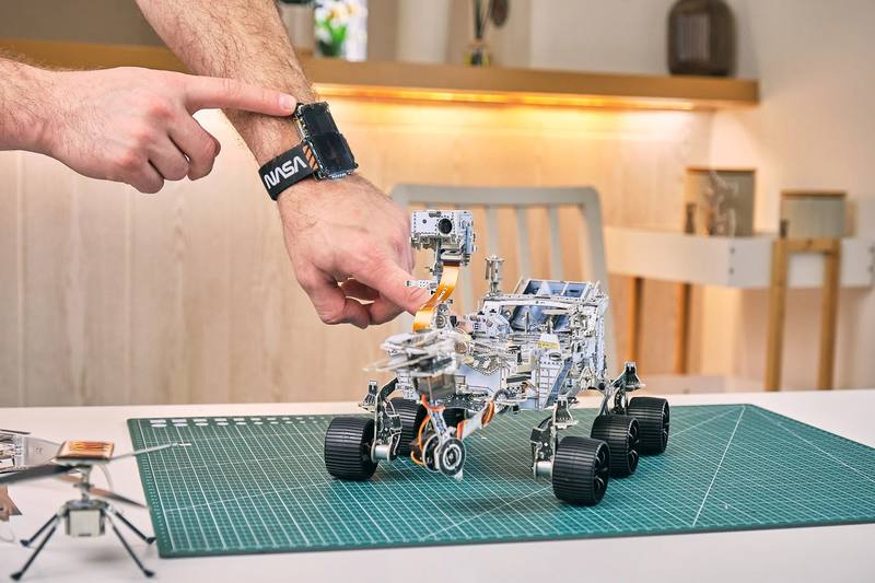 A wrist wearing the NASA Artemis Watch reaches in to adjust the assembled Perseverance rover kit, a model of the Ingenuity helicopter in the foreground.